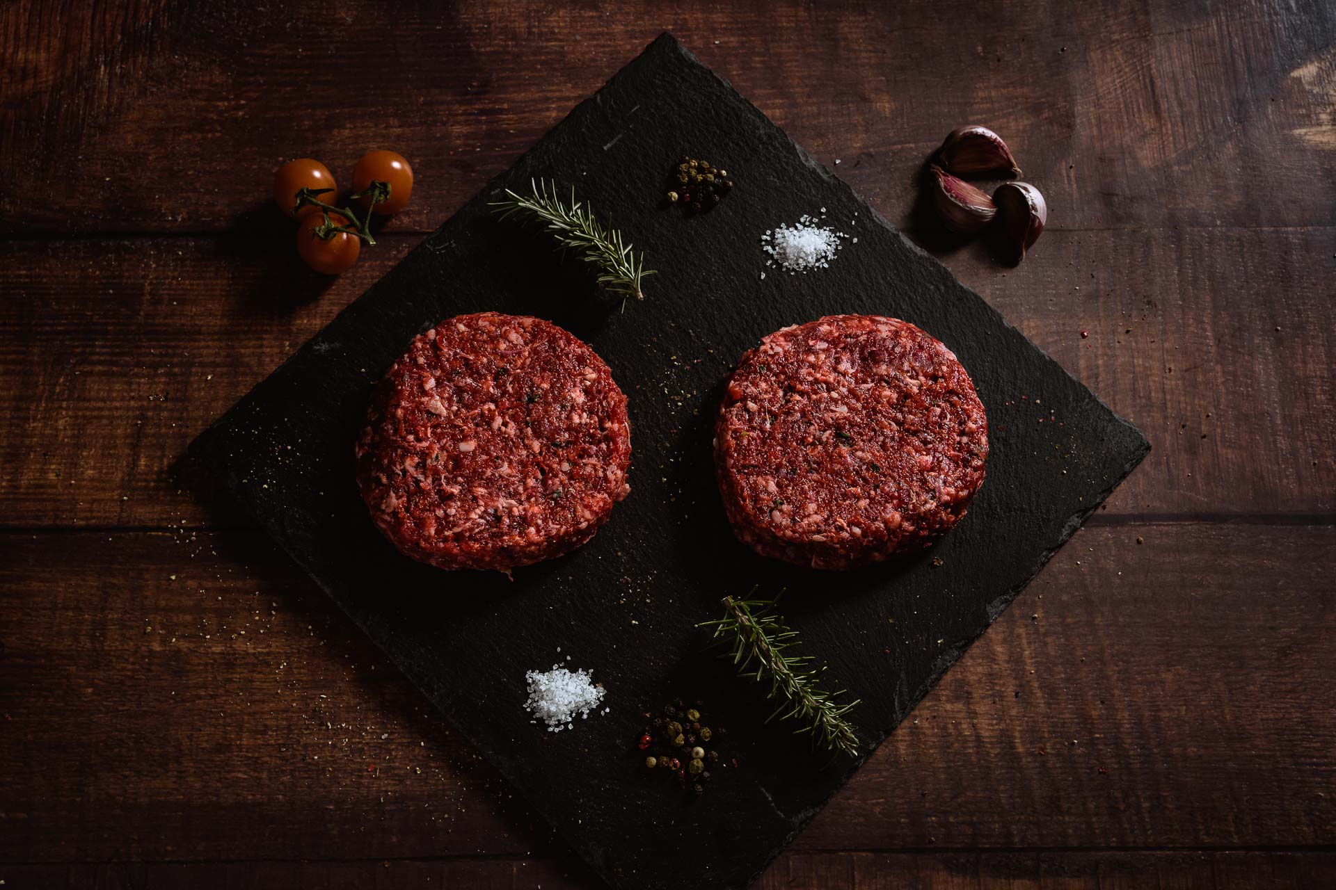 Dos hamburguesas de vaca tudanca crudas sobre pizarra negra con tomates cherry, romero, sal y ajos